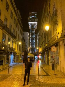 A imagem mostra uma rua de Lisboa à noite, com iluminação amarela suave proveniente de postes de luz. Ao fundo, destaca-se o famoso Elevador de Santa Justa, iluminado e imponente, com sua estrutura metálica detalhada e elementos neogóticos. A rua é pavimentada com pedras portuguesas e cercada por edifícios tradicionais com varandas de ferro forjado, típicos da arquitetura lisboeta.No centro da cena, em primeiro plano, uma pessoa está posando de pé, usando roupas escuras. O ambiente parece tranquilo, com poucas pessoas visíveis, e o reflexo da iluminação no chão molhado sugere que pode ter chovido recentemente. A composição transmite um clima noturno e sereno, realçando o charme histórico da cidade.